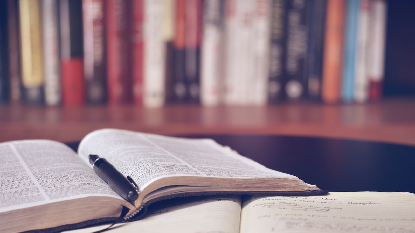 A stack of several books on a wooden table next to a pair of reading glasses
