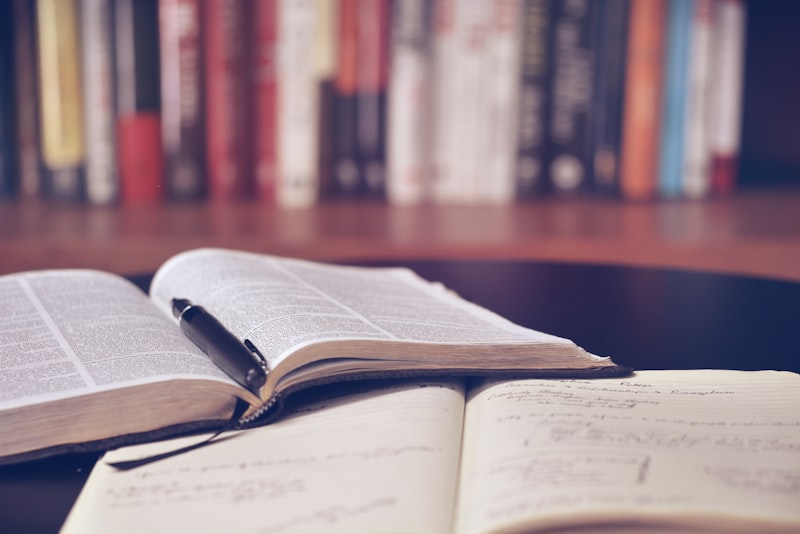 A stack of several books on a wooden table next to a pair of reading glasses